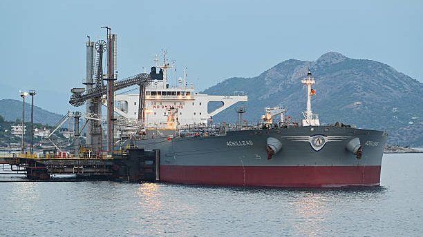 Barriles de petróleo crudo apilados en un puerto con un barco tanquero al fondo